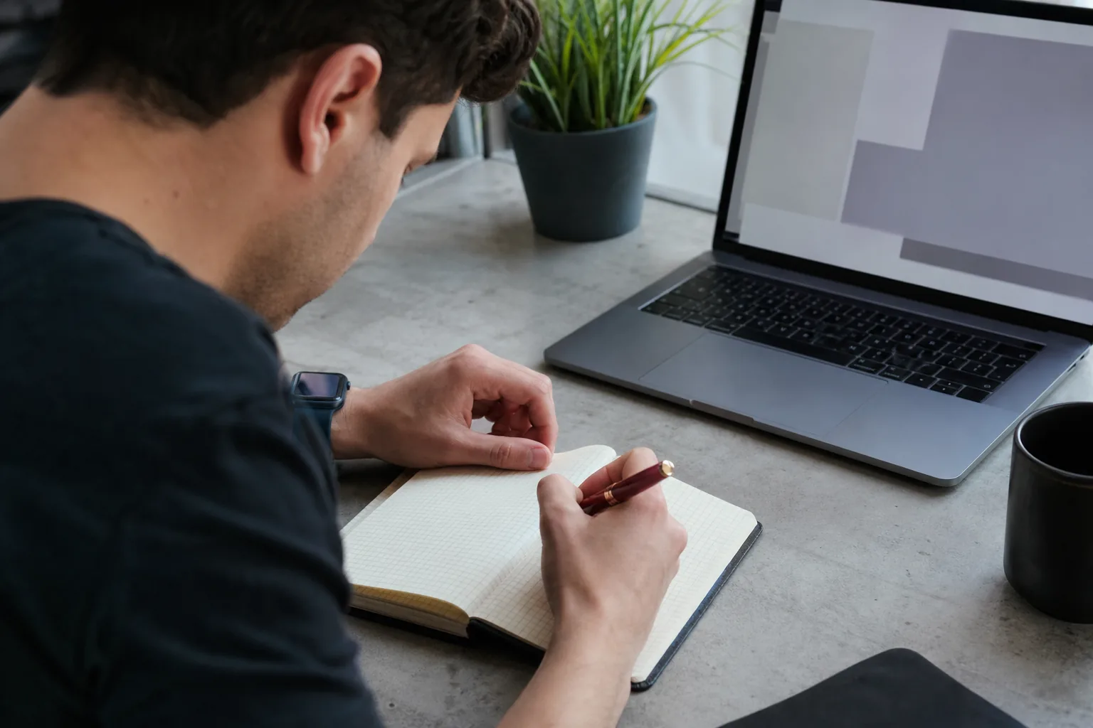 Team member writing a scheduled job in plain words in a notebook next to a laptop