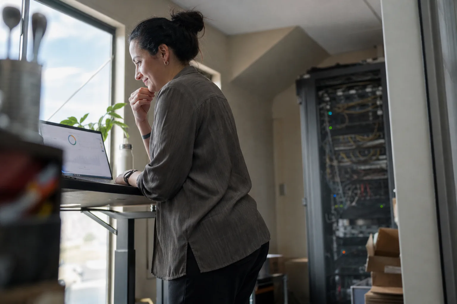 Operations manager checking scheduled agent results on a laptop, with an old self hosted server rack in the corner